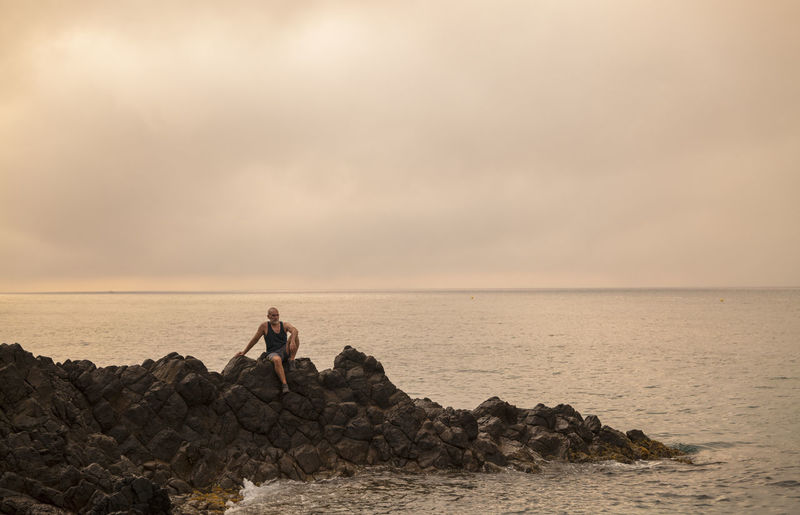 Man sitting on rock looking at scenic view of beach against sky
