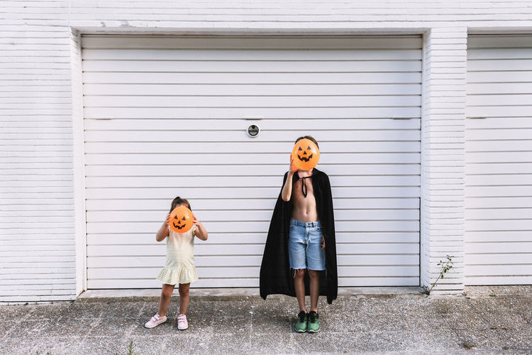 Boy and girl covering faces with balloons in shape of scary pumpkin while standing together on street during daytime