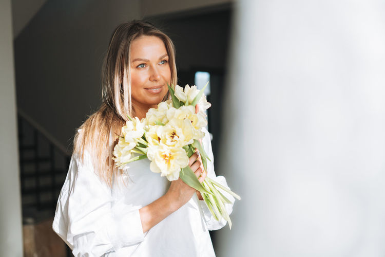 Young woman with blonde long hair in shirt with bouquet of flowers in hands near window at home