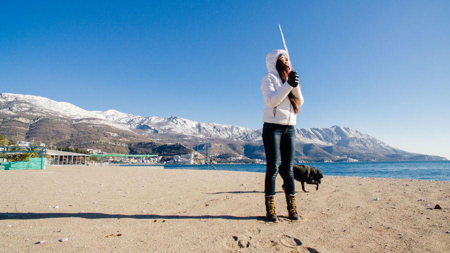 Woman holding icicle while standing with dog at beach against sky