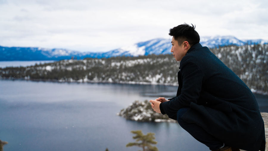 Side view of young man crouching over lake against sky