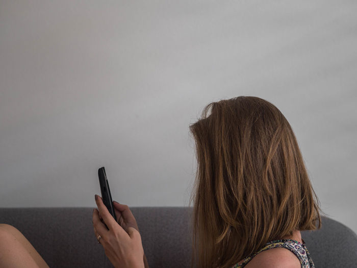 Side view of woman using phone on sofa against white wall