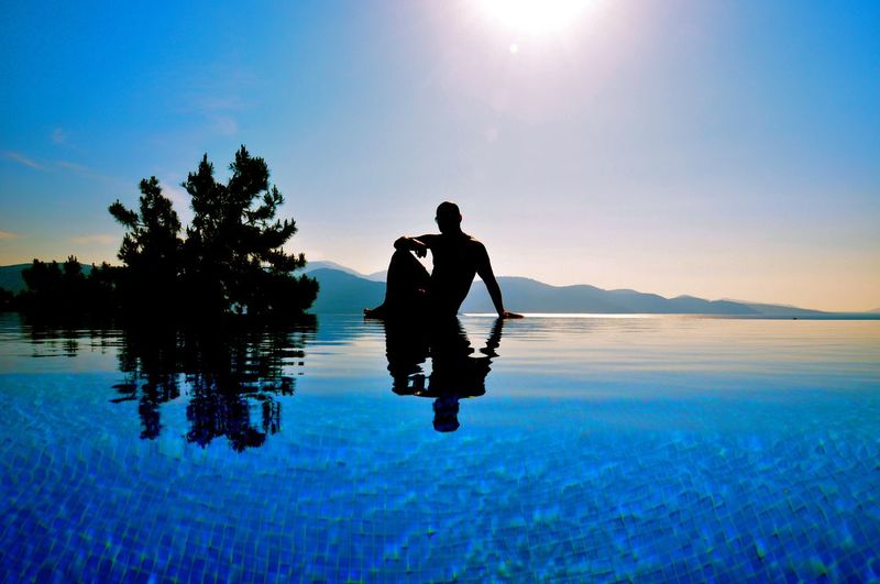 Silhouette man sitting at infinity pool against sky on sunny day