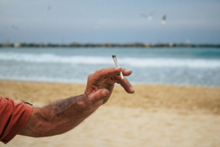 Cropped image of man holding cigarette at beach against sky