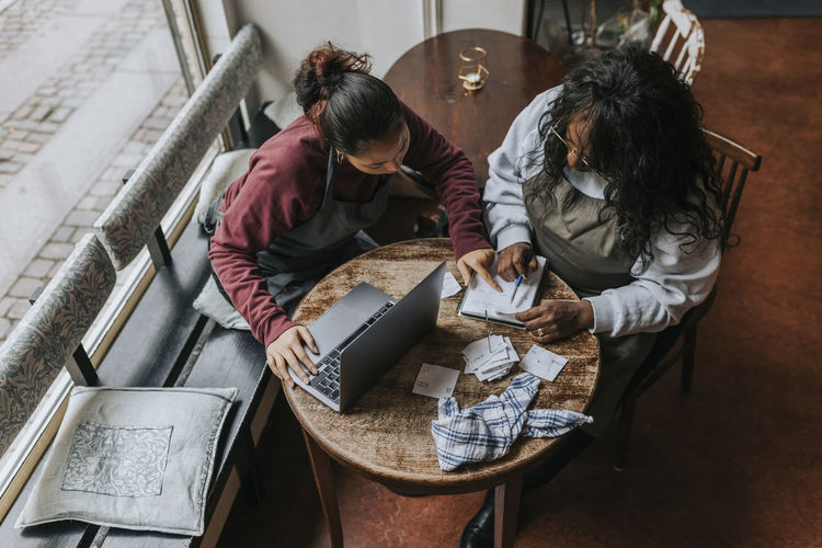 High angle view of female cafe owners discussing over diary while sitting at table in coffee chop