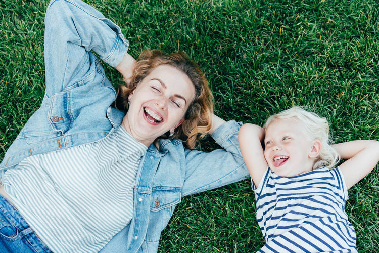 Top view of carefree laughing mother and daughter lying on green grass and enjoying summer
