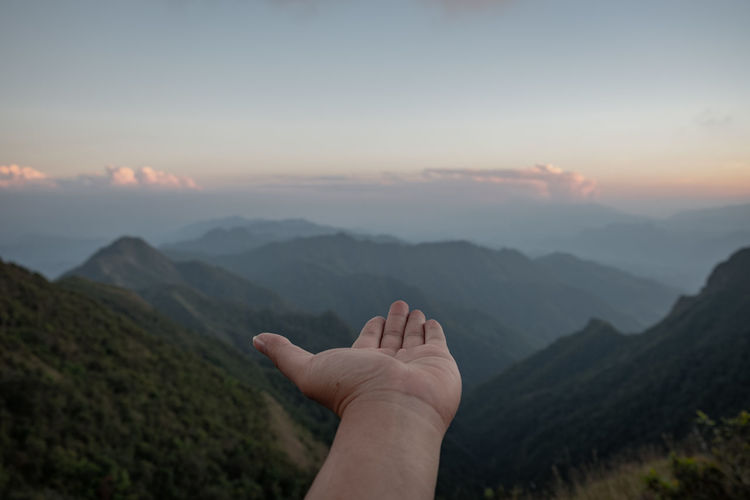 Cropped hand of woman reaching mountain against sky during sunset