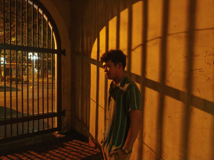 Side view of young man standing against wall and gate