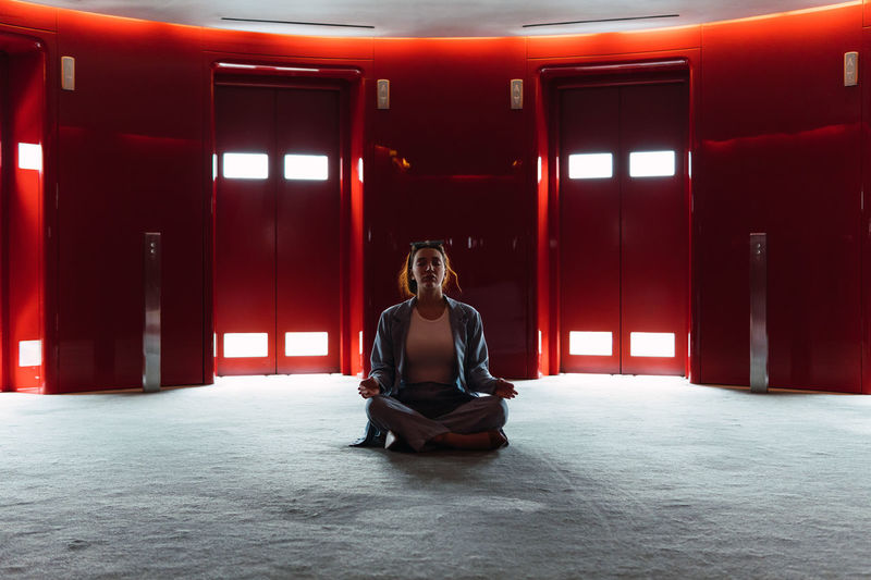 Full body of peaceful female sitting in lotus pose while meditating in spacious foyer with red elevators with glowing lights