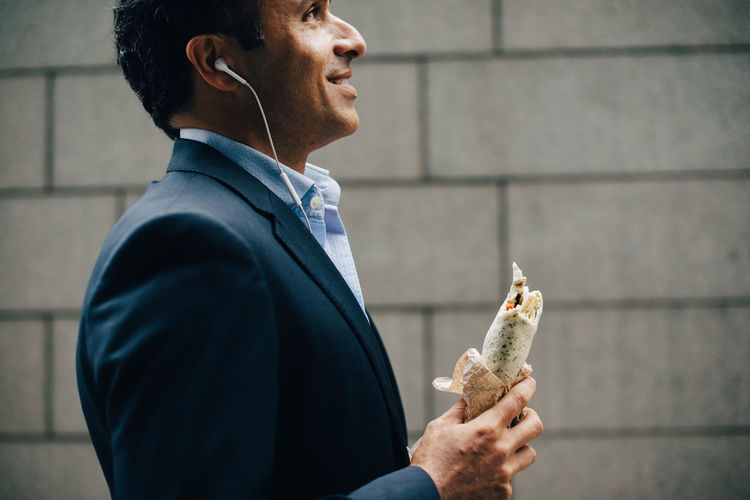 Side view of businessman listening from in-ear headphones while holding wrap sandwich against wall in city