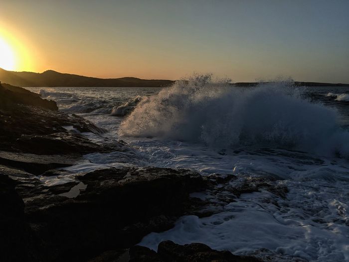 Waves splashing on rocks against sky during sunset