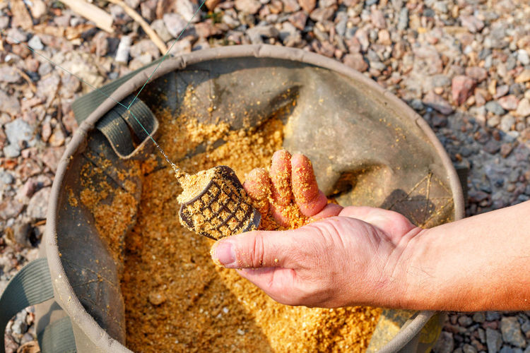 A metal feeder for fish in the hand of the fisherman there is with bait against of tarpaulin bucket.