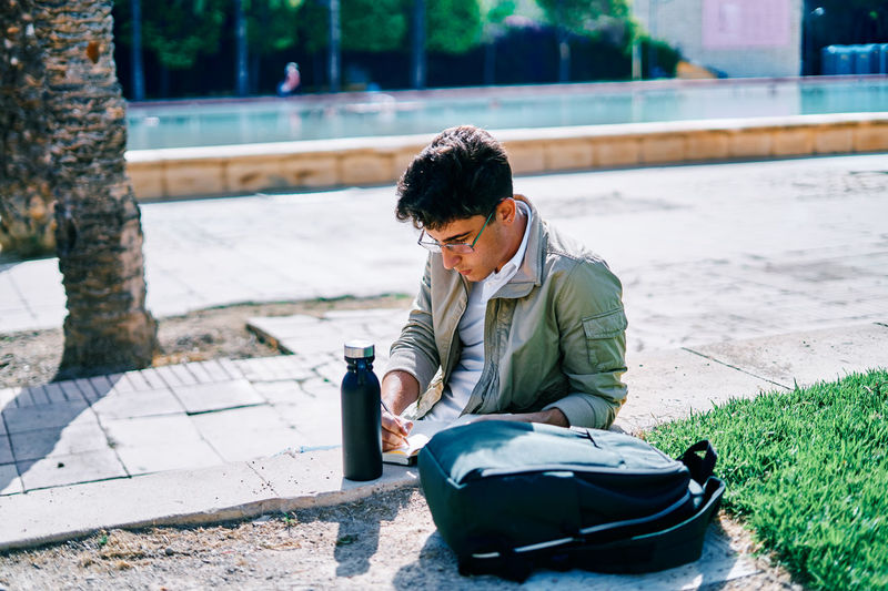 Focused male student sitting on stone bench in university campus and writing in notepad while doing homework on sunny day