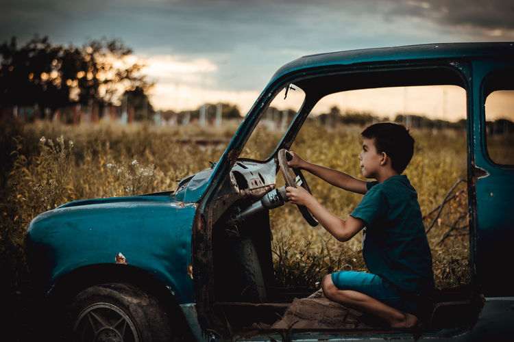 Side view of boy crouching in broken vintage car on field