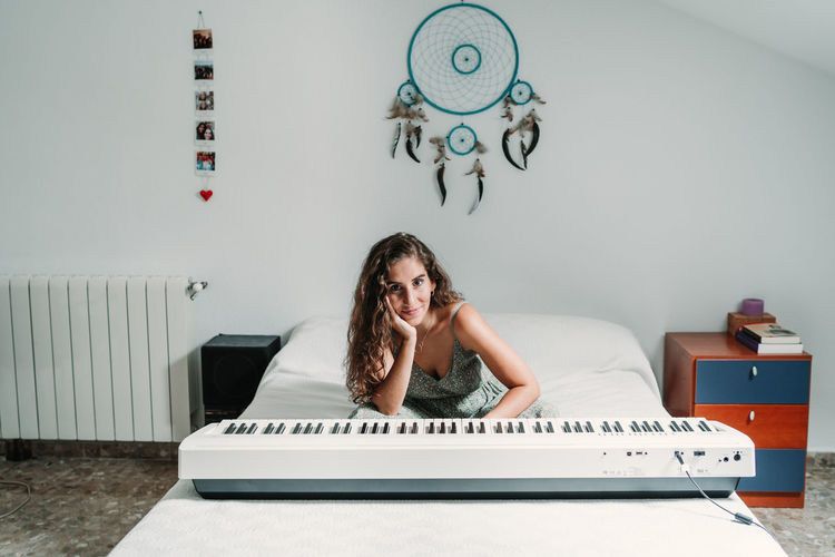 Calm ethnic female musician sitting on bed with contemporary electric piano and looking at camera while leaning on hand