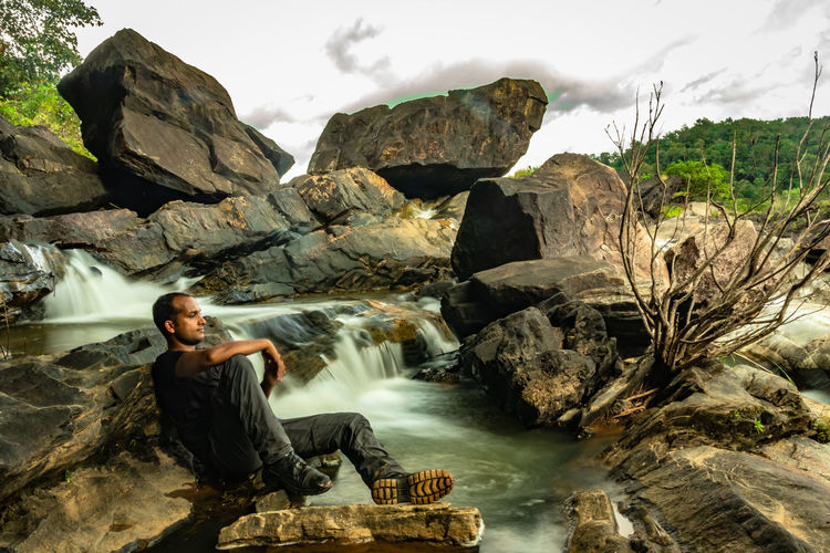Young man sitting on rock against waterfall