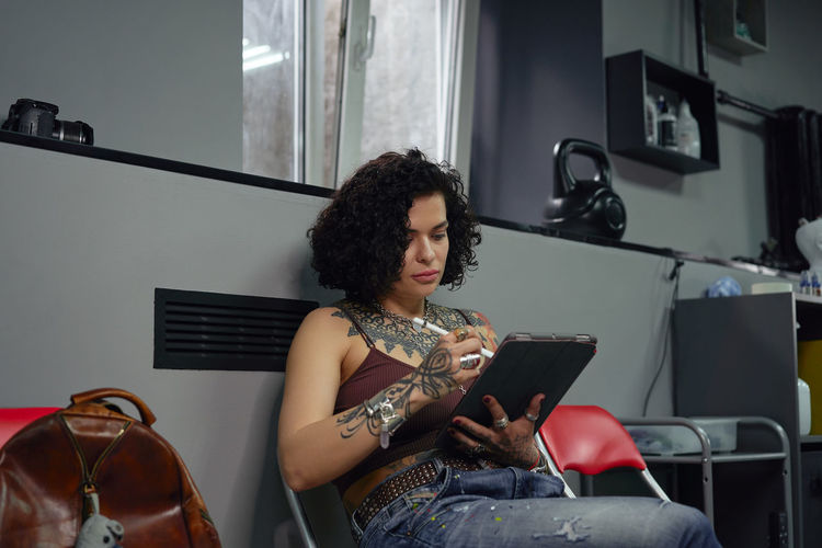 Concentrated tattoo master in casual clothes sitting on chair with tablet and pen while drawing tattoo sketches in light tattoo salon
