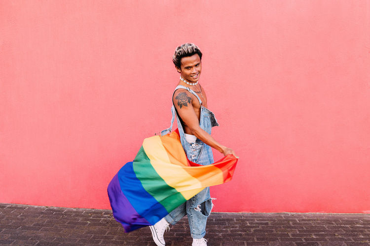 Portrait of young man holding rainbow flag in front of pink wall