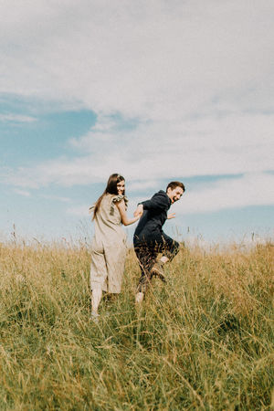 Young couple holding hands while walking on grassy field against cloudy sky