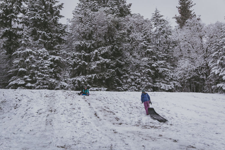 Rear view of caucasian girl walking up on snow covered land with hispanic girl sledding down hill