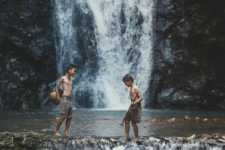 Smiling shirtless boys standing by waterfall