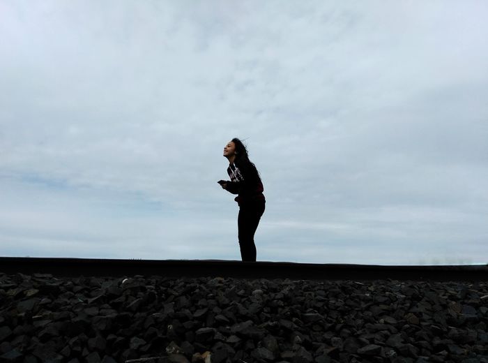 Happy teenage girl standing at railroad track against cloudy sky