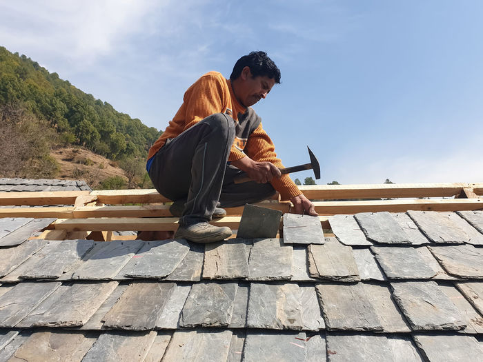Low angle side view of a villager man laying roof slate tiles on rooftop of building