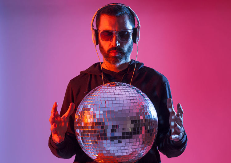 Studio portrait of a bearded deejay with headphones and sunglasses against red and blues background.