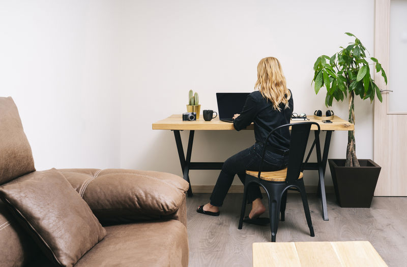 Beautiful blonde caucasian woman works from her living room with her laptop on a wooden desk. she wears black casual clothes.