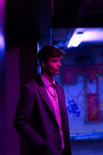 Young man looking away while standing against illuminated wall