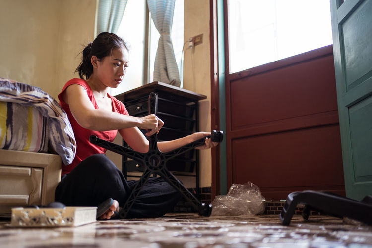 Side view of ethnic asian female screwing wheels to chair base while sitting cross legged on floor near bed and assembling furniture in cozy bedroom at home