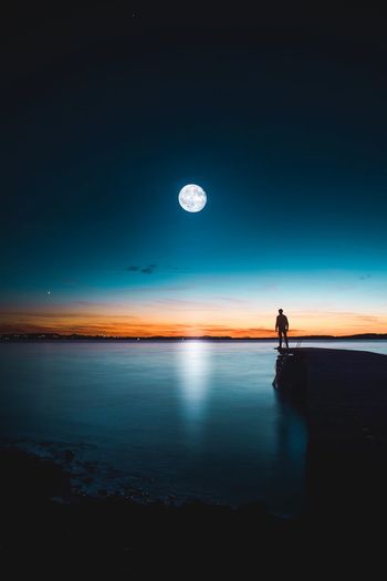 Silhouette man standing at lakeshore against moon at dusk