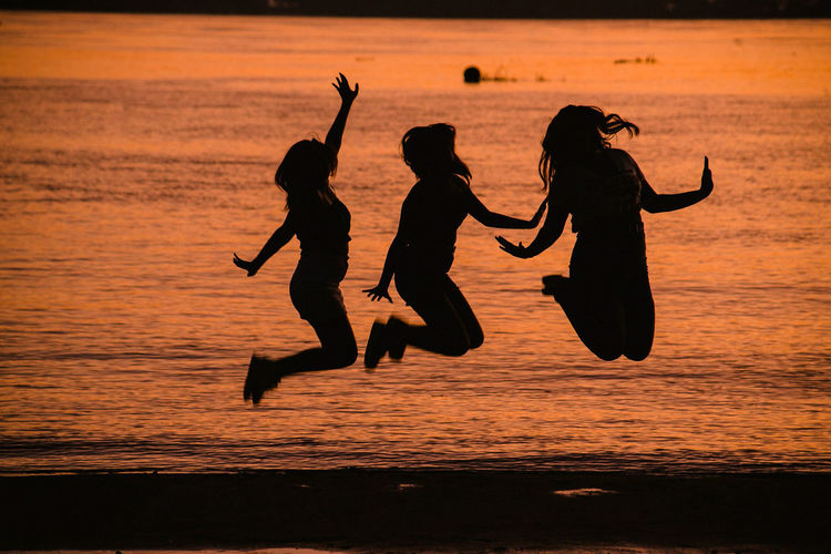 Silhouette people jumping on sea against sky during sunset