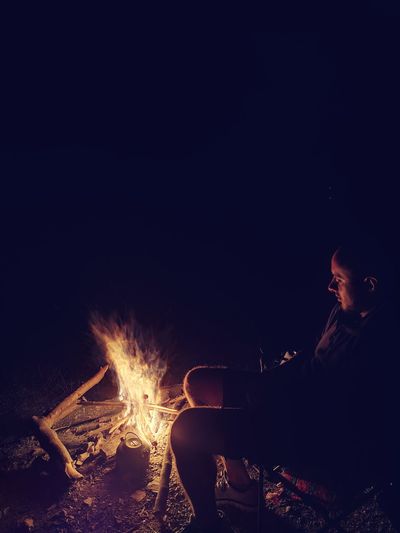 Side view of man sitting by campfire in dark at night