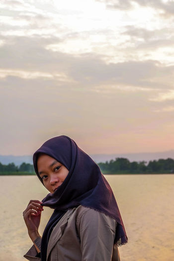 Portrait of woman standing in sea against sky during sunset wearing hijab