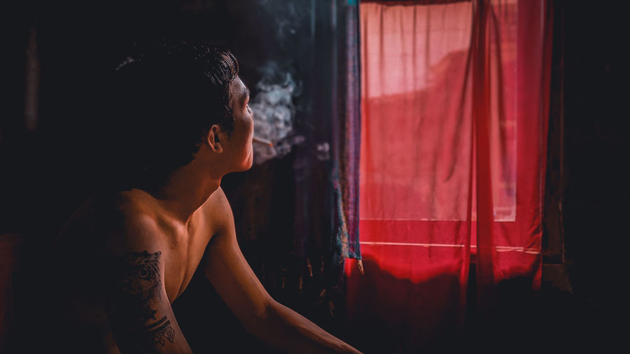 Shirtless man smoking cigarette while looking at window
