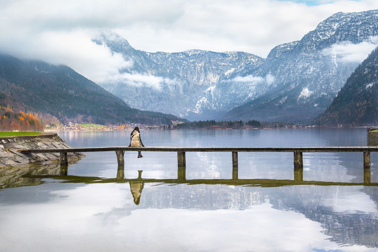 Rear view of woman sitting on bridge over hallstatter see against mountains in hallstatt