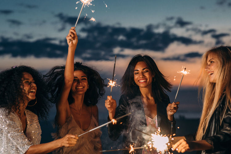 Cheerful friends holding sparklers while standing against sky