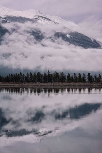 Scenic view of mountain, trees, and low clouds reflected in lake 