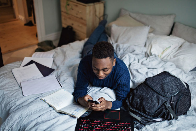 High angle view of distracted teenage boy using mobile phone while lying on bed by books at home
