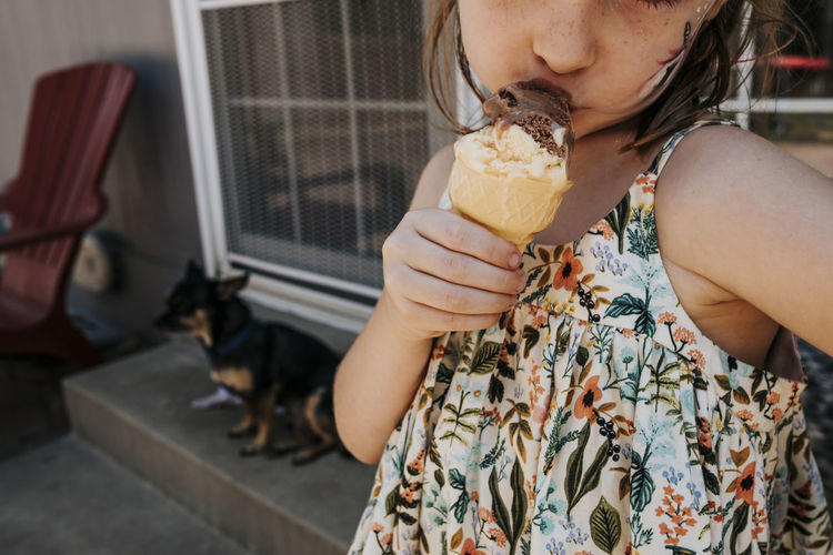 Midsection of cute girl eating ice cream cone while standing against house