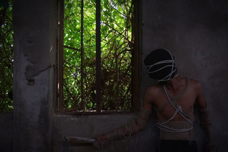 Shirtless man with ropes tied on body standing by wall in forest