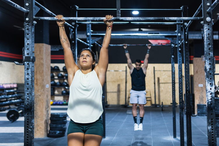 Woman in activewear hanging on metal crossbars during intense training with strong muscular unrecognizable man in light modern gym