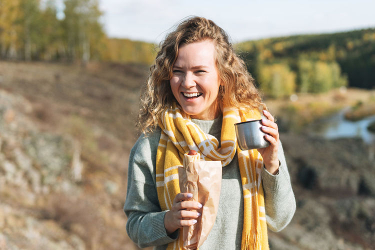Young happy beautiful woman traveller with curly hair eating hot dog and drinking tea on nature view 