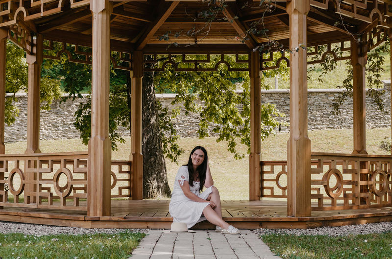Portrait of beautiful young woman wearing white dress sitting under wooden pavilion in park.