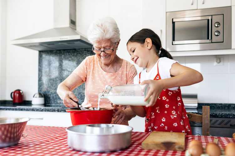 Grandmother in eyewear with whisk near granddaughter with bottle of milk cooking dough at table in house