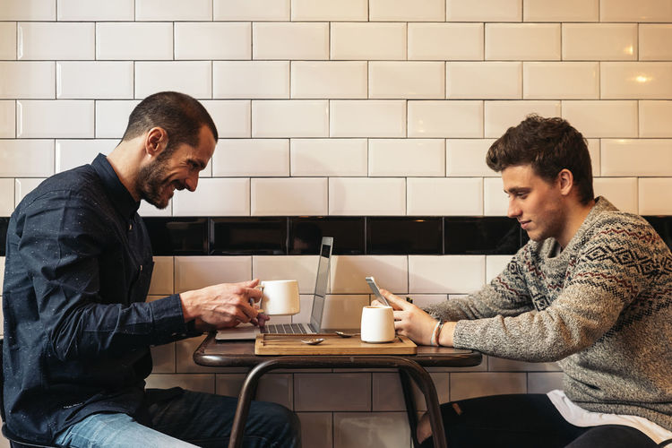 Colleagues with coffee cups using technologies while sitting at table in cafe