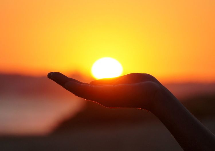 Optical illusion of hand holding sun against sky during sunset