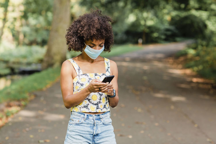 Mid adult woman wearing face mask using mobile phone while standing on road at park