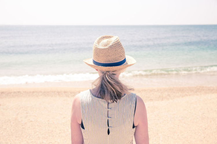 Rear view of mature woman wearing hat while standing at beach against clear sky during sunny day